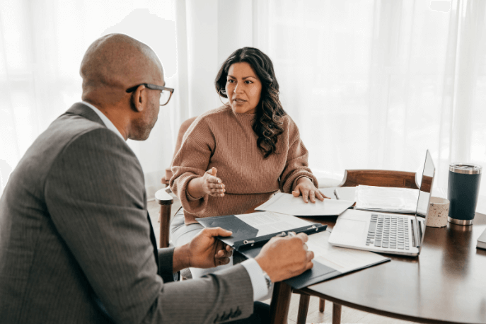 Two people talking at a table. Laptop and papers on table.