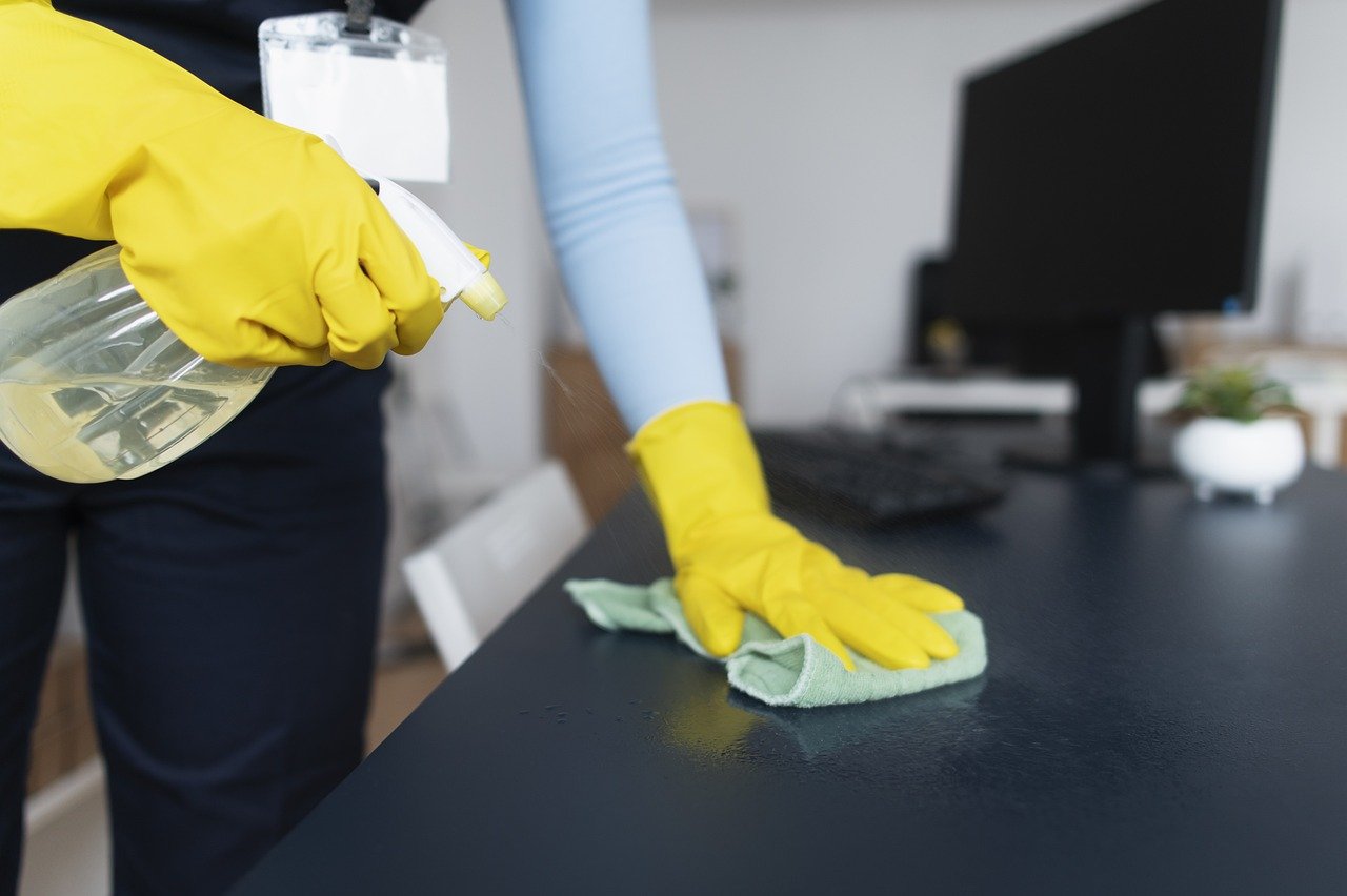 Person cleaning an office desk, wearing yellow gloves. Image by Pixabay