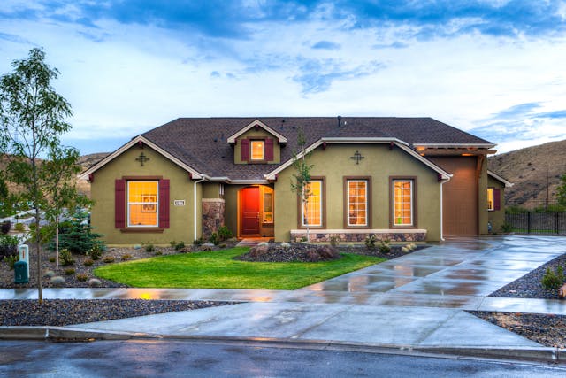 Beigh house, lights on, wet street and driveway. Image by Pexels