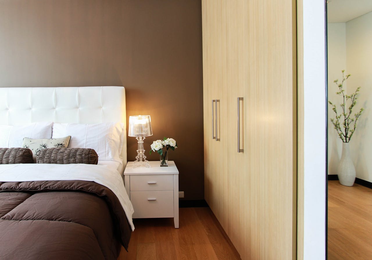 White bed with brown bedding nect to a white night table. Image by Pexels