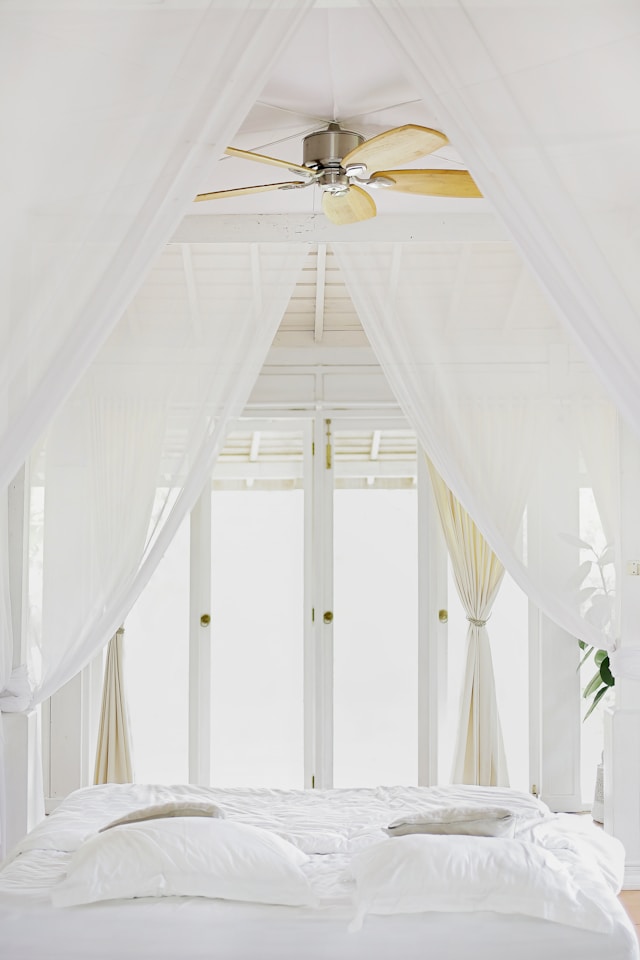 A bedroom with white bedding and white curtains, windows. Image by Unsplash
