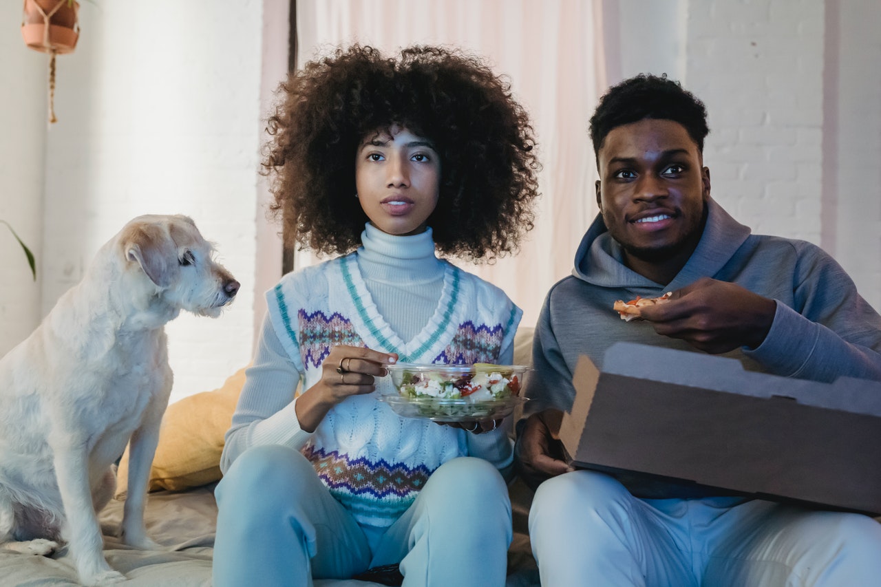 A couple sitting on a bed eating, a white dog is watching