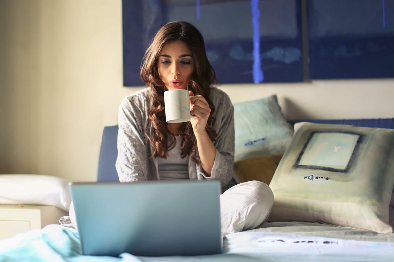 Person blowing on a cup of coffee, laptop on a bed. Image by Pexels