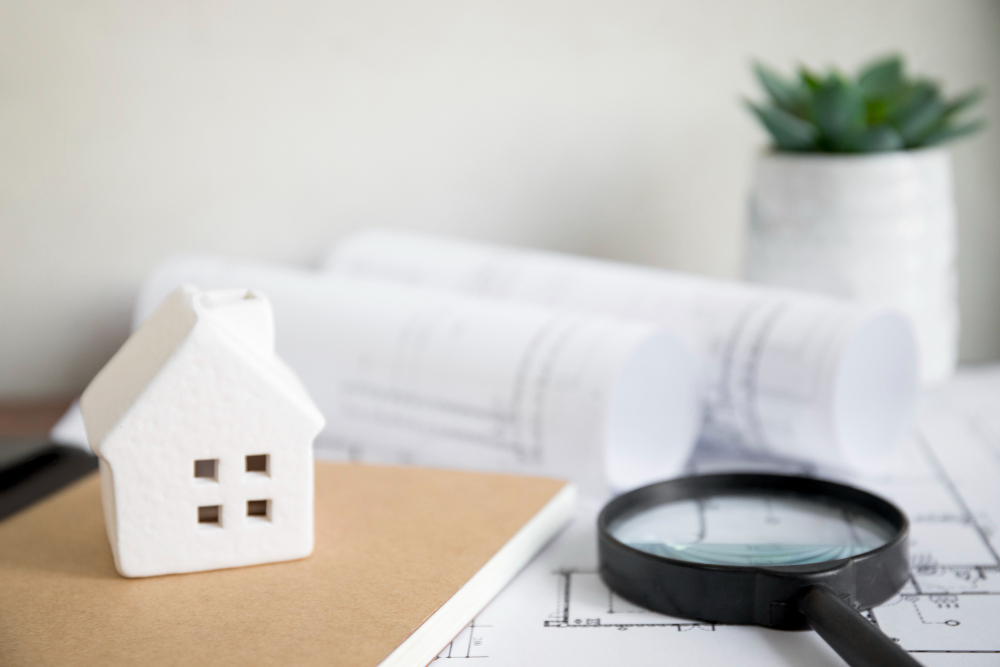 small white house, magnifying glass, plant and paperwork on a table. Image by by Freepik