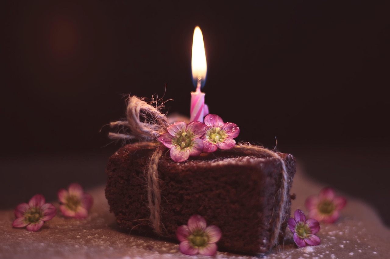 Brownie wrapped as a present, with lit birthday candle on top. Lots of pink flowers. Image by Pixabay