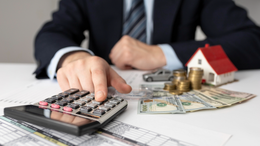 Person it a suit, using a calculator, money, car and house on a table.