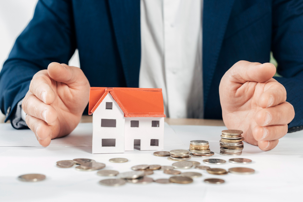 Hands surrounded by a small house and coins on a table.