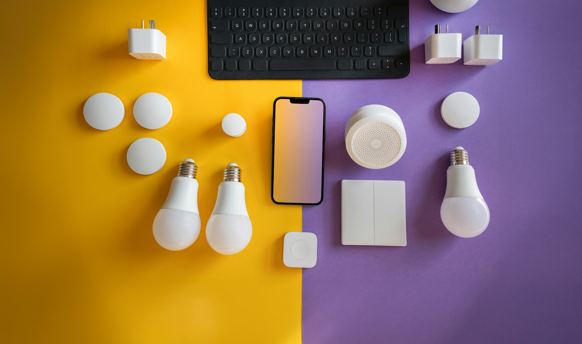 Computer keyboard, light bulbs, and other electronics on a purple and yellow background representing energy efficient lighting. Image via Unsplash