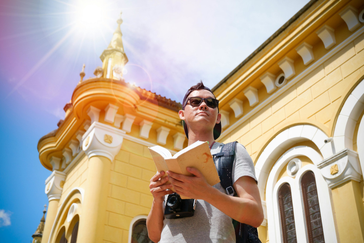 Person with sunglasses and a hat, reading a book outside in front of a yellow and white building. Image by Pexels.