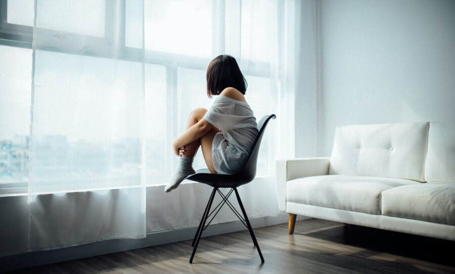 Person siting with on a chair holding her legs looking out a window. Sofa in the background.